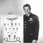 Achievement Captain Audie Murphy in uniform standing next to a framed display case of the medals he earned during World War II.  of Audie Murphy